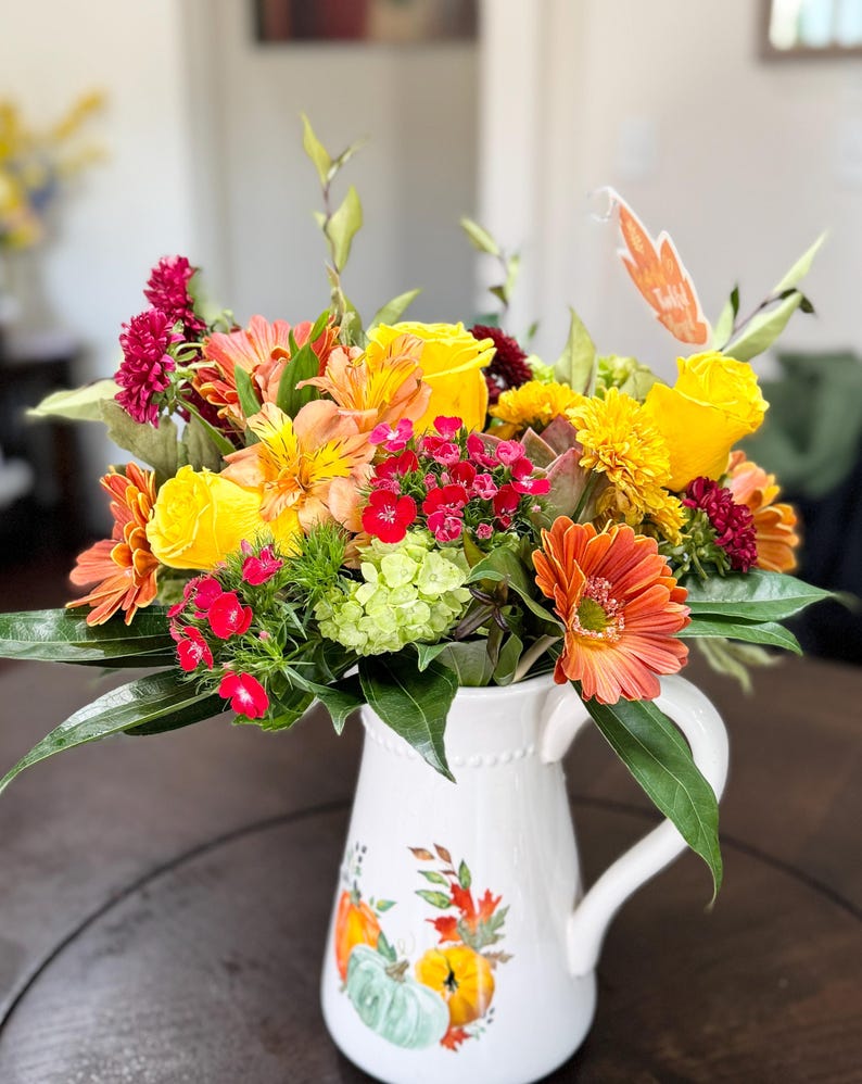 May include: A bouquet of yellow, orange and red flowers in a white jug with a pumpkin design. The flowers are arranged in a vase with a white background.