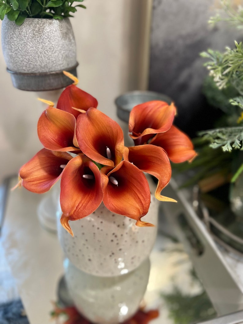 May include: A bouquet of orange calla lilies in a speckled white ceramic vase. The flowers have a rich, warm color with a hint of yellow at the tips. The vase is round and has a textured surface, adding a rustic touch to the floral arrangement.