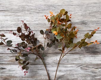 May include: Two sprigs of artificial eucalyptus with green leaves and small pink and orange flowers. The sprigs are arranged on a light gray wooden background.