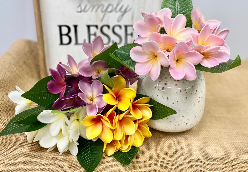 May include: A bouquet of artificial flowers in a white ceramic vase. The flowers are pink, purple, white, and yellow. The vase has a speckled pattern. The flowers are arranged in a vase with a sign that says "Simply Blessed" in the background.