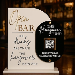 Könnte beinhalten: Ein weißes und schwarzes Schild für eine offene Bar mit goldfarbenen Buchstaben. Auf dem Schild steht "Open Bar, Die Getränke gehen auf uns! Der Kater geht auf dich!" und "The Honeymoon Fund". Enthält einen QR-Code.