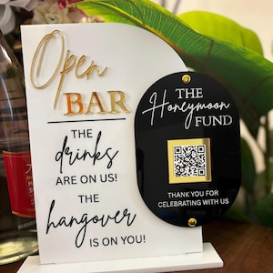May include: A white and black open bar sign with gold lettering. The sign reads "Open Bar" and "The drinks are on us! The hangover is on you!" A black sign with a QR code reads "The Honeymoon Fund."