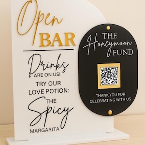 May include: A white and black open bar sign with gold lettering. The sign reads "Open Bar," "Drinks are on us!" and "Try our love potion: The Spicy Margarita." A black sign with "The Honeymoon Fund" and a QR code is attached.