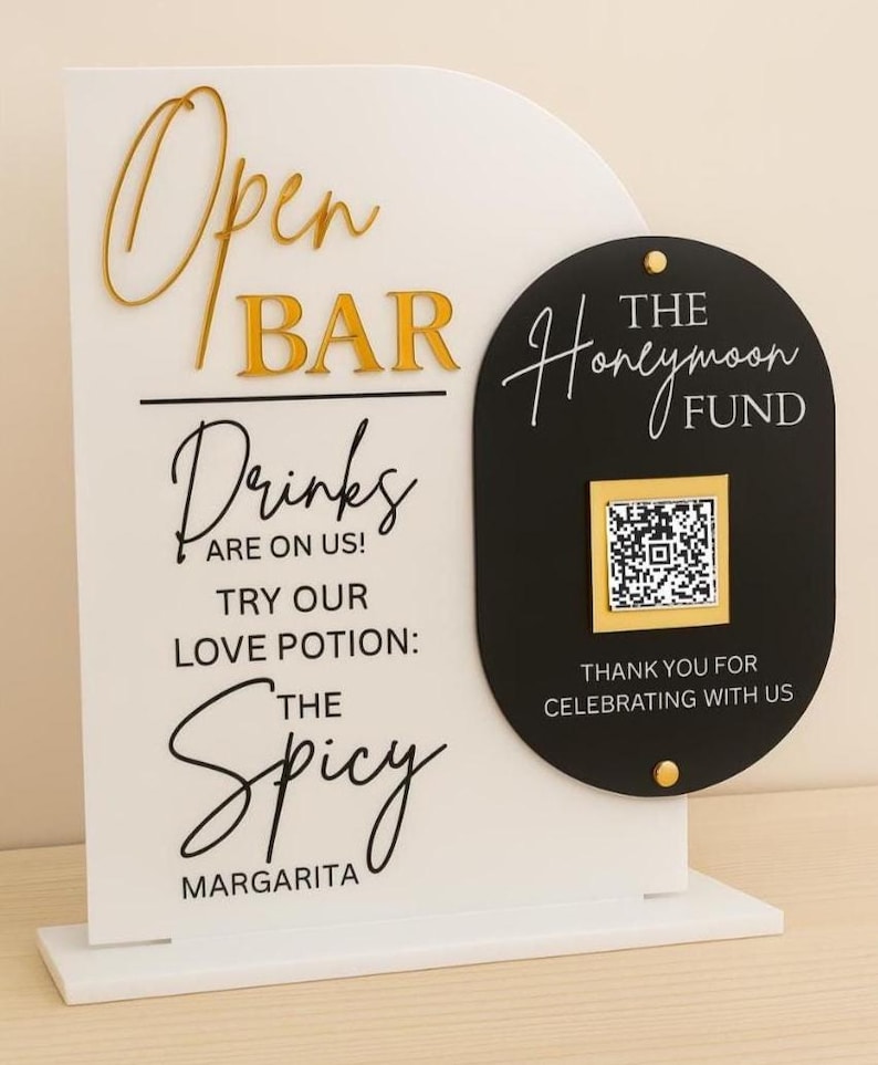 May include: A white and black open bar sign with gold lettering. The sign reads "Open Bar," "Drinks are on us!" and "Try our love potion: The Spicy Margarita." A black sign with "The Honeymoon Fund" and a QR code is attached.