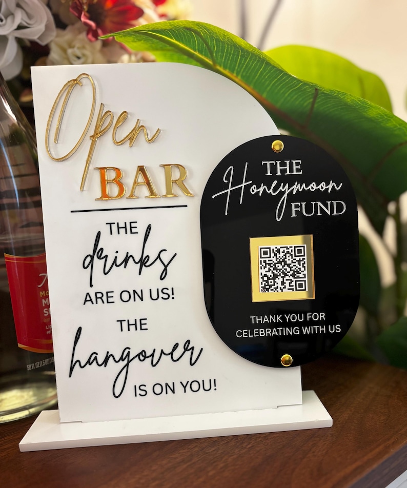 May include: A white and black open bar sign with gold lettering. The sign reads "Open Bar" and "The drinks are on us! The hangover is on you!" A black sign with a QR code reads "The Honeymoon Fund."