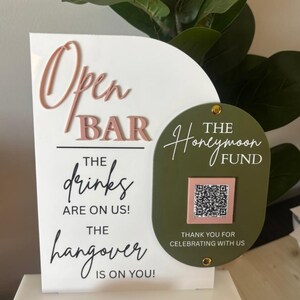 May include: A white sign with the words "Open Bar" in pink and "The drinks are on us! The hangover is on you!" in black. A green oval with "The Honeymoon Fund" and a QR code is attached.