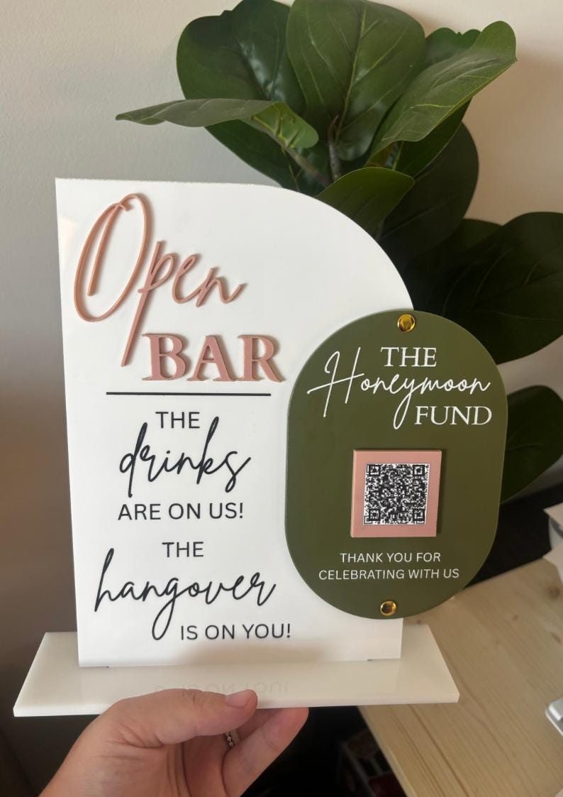 May include: A white sign with the words "Open Bar" in pink and "The drinks are on us! The hangover is on you!" in black. A green oval with "The Honeymoon Fund" and a QR code is attached.