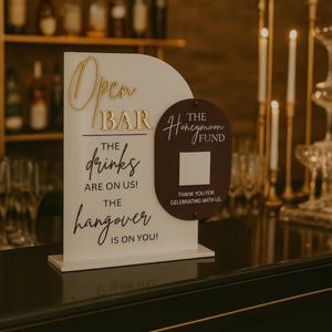 May include: A white and brown wedding sign with gold lettering that reads "Open Bar" and "The Honeymoon Fund." The sign also says "The drinks are on us! The hangover is on you!" and "Thank you for celebrating with us."