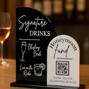 May include: A black and white acrylic sign set on a black base. The taller black section features the text Signature DRINKS, Blushing Bride, and Love on the Rocks with drink icons. The shorter white section displays Honeymoon fund, a QR code, and text.