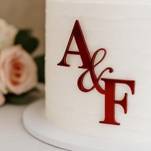 Może przedstawiać: Biały tort oblany lukrem z czerwonym monogramem z literami "A & F". Ciasto stoi na białym stojaku na ciasto, z rozmytym kwiatowym tłem. Czerwone litery są w czcionce skryptowej.