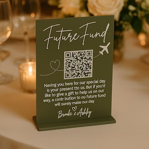 May include: A green table sign with the words "Future Fund" in white script. It includes a QR code, a heart, and a small airplane graphic. The sign's message is about a future fund. The names "Brooke & Ashky" are at the bottom.