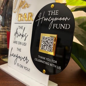 May include: A white and black sign with gold accents. The sign reads "Open Bar" and "The drinks are on us! The hangover is on you!" A black sign next to it says "The Honeymoon Fund" with a QR code.