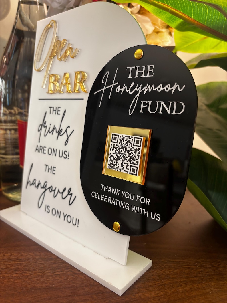 May include: A white and black sign with gold accents. The sign reads "Open Bar" and "The drinks are on us! The hangover is on you!" A black sign next to it says "The Honeymoon Fund" with a QR code.