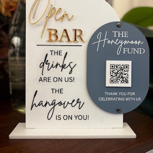 May include: A white acrylic bar sign with gold lettering that reads "Open BAR". Below, it says "The drinks are on us! The hangover is on you!" A blue oval has "The Honeymoon Fund" and a QR code.