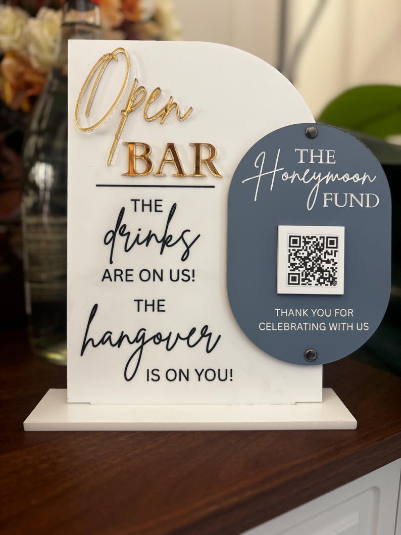 May include: A white acrylic bar sign with gold lettering that reads "Open BAR". Below, it says "The drinks are on us! The hangover is on you!" A blue oval has "The Honeymoon Fund" and a QR code.