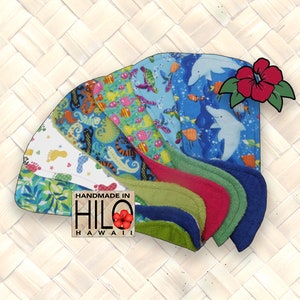 May include: A set of seven colorful dishcloths with various tropical and ocean-themed patterns. The dishcloths are made of a soft, absorbent material and are handmade in Hilo, Hawaii. The dishcloths are arranged in a fan shape with a red hibiscus flower on top.