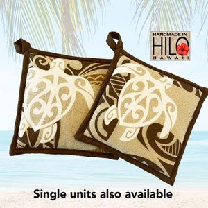 May include: Two square pot holders with a brown border and a beige background featuring a white turtle design. The pot holders have a loop for hanging. The text "HANDMADE IN HILO HAWAII" is visible.