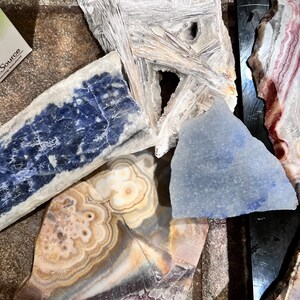 Pu&ograve; includere: Una collezione di cinque tipi diversi di pietre levigate, tra cui una bianca, una blu, una blu chiaro, una marrone e bianca e una rosa e bianca. Le pietre sono disposte su una superficie scura.