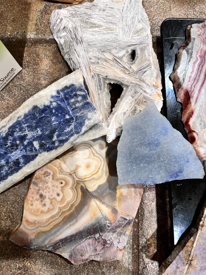 Pu&ograve; includere: Una collezione di cinque tipi diversi di pietre levigate, tra cui una bianca, una blu, una blu chiaro, una marrone e bianca e una rosa e bianca. Le pietre sono disposte su una superficie scura.