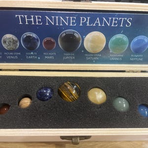 May include: A wooden box displaying a set of nine gemstone spheres representing the planets. The box lid features the text "THE NINE PLANETS" above the spheres, with each planet labeled. The spheres are various colors and textures, including clear quartz, tiger eye, and aventurine.