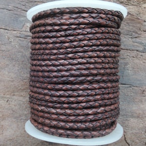 Peut inclure: Une bobine de cordon en cuir marron, tressé en forme de losange. Le cordon est enroulé serré autour d'une bobine en plastique blanc.