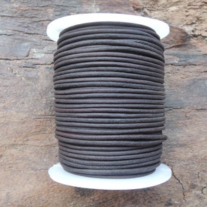 Può includere: Una bobina di cordoncino in pelle marrone scuro, avvolta strettamente e presentata su uno sfondo di legno rustico. Il cordoncino è rotondo e ha una superficie strutturata, ideale per la creazione di gioielli o progetti artigianali.