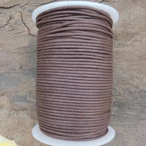 Può includere: Una bobina di cordoncino di cotone cerato marrone. Il cordoncino è avvolto strettamente attorno a una bobina di plastica bianca. Il cordoncino è di colore marrone medio uniforme. Lo sfondo è una superficie di legno naturale.