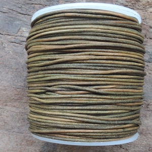 May include: A spool of brown and green leather cord. The cord is wound tightly around a white plastic spool.