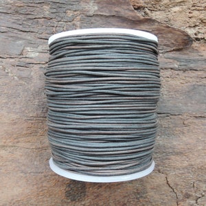 Puede incluir: Un carrete de cordón de cuero gris oscuro, enrollado firmemente alrededor de un carrete de plástico blanco. El cordón es liso y parece ser flexible.