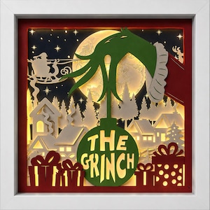 May include: A layered paper shadow box art piece in a white frame featuring a green hand holding a green ornament with the text THE GRINCH. The scene includes a backlit moon, a silhouette of a sleigh with reindeer, houses, and red gift boxes.