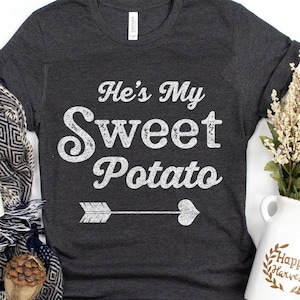 May include: A dark grey t-shirt with a white graphic that says "He's My Sweet Potato" with an arrow pointing to the right and a heart at the end of the arrow.