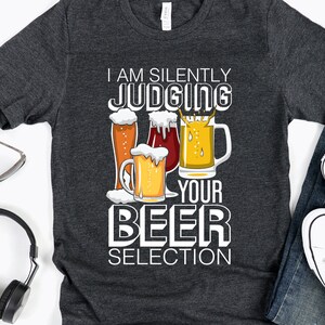 May include: A dark gray t-shirt with the text "I am silently judging your beer selection" and a graphic of four beer glasses.