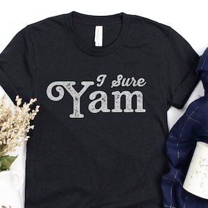 May include: A black t-shirt with the text "I Sure Yam" in white, displayed on a white surface with a white vase of dried flowers, a blue and white scarf, a white mug, and a pine cone.