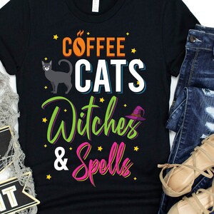 Puede incluir: Camiseta negra con un gráfico de temática de Halloween que dice "Coffee Cats Witches & Spells" en texto colorido con estrellas y una ilustración de un gato negro.