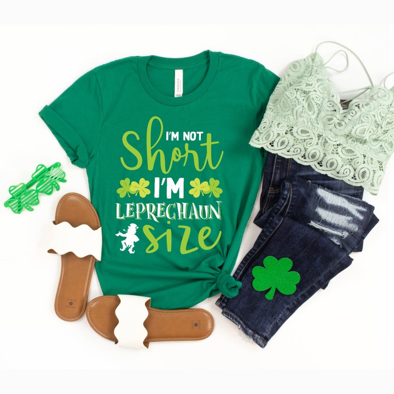 Pu&ograve; includere: Maglietta verde con il testo "I'm not short I'm Leprechaun size" e grafiche a trifoglio. La maglietta &egrave; abbinata a jeans blu, un top di pizzo verde e sandali bianchi.