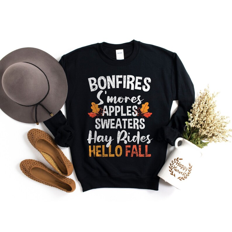 Puede incluir: Sudadera negra con un estampado gr&aacute;fico blanco que dice "Bonfires, S'mores, Apples, Sweaters, Hay Rides, Hello Fall".