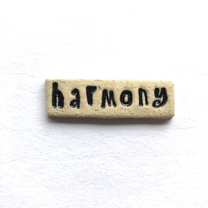 May include: A rectangular, beige ceramic tile with the word "harmony" in black, handwritten-style lettering. The tile has a textured surface and is set against a white background.