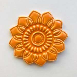 May include: A ceramic tile featuring a raised, orange flower design. The flower has a circular centre with a smaller circle in the middle.