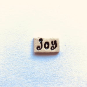 May include: A small, rectangular tile with the word "Joy" written in black cursive lettering.