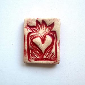 May include: A rectangular, off-white ceramic tile with a red heart and flame design. The heart is in the center, with a flame-like shape above it. The tile has a slightly rough texture and is set against a white background.