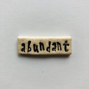 Pode incluir: Um pequeno azulejo retangular de cerâmica de cor creme com a palavra "abundante" escrita em letras pretas.