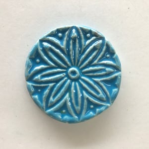 Puede incluir: Un azulejo de cerámica redondo, azul, con un diseño floral blanco en relieve. El azulejo tiene un acabado brillante.