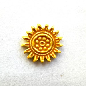 May include: A yellow, sun-shaped ceramic bead with a detailed design. The bead features a central circle with smaller circles and radiating points around the edge. The bead is set against a white background.