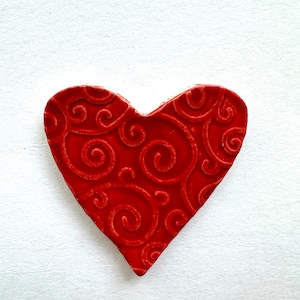 May include: A red heart-shaped ornament with a textured surface and a swirling pattern. The heart is a solid, vibrant red colour and is set against a white background. The design features a repeating, elegant scroll motif.