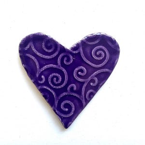 May include: A heart-shaped object with a glossy deep purple finish featuring a raised, lighter purple swirling vine pattern across the surface. The edges of the heart are smooth and rounded, creating a distinct contrast between the dark base colour and the intricate, repeating scrollwork design that covers the entire front face of the piece.
