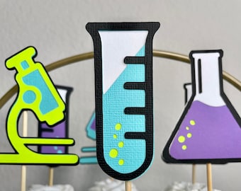 May include: A set of four science-themed cupcake toppers. The toppers are made of paper and feature a beaker, a test tube, a microscope, and a flask. The toppers are in various colors, including green, blue, purple, and yellow.
