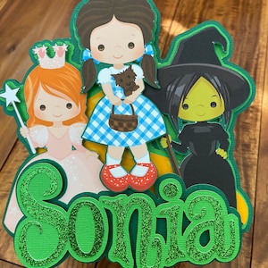 May include: A green and yellow cake topper featuring Dorothy from the Wizard of Oz, the Wicked Witch of the West, and a fairy. The topper has the name "Sonia" written in green glitter.