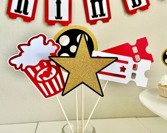 May include: A collection of movie-themed party decorations. Includes a gold glitter star, popcorn, film reel, and ticket cutouts on wooden sticks, displayed in a glass jar. A banner spelling "BIRTHDAY" hangs in the background.