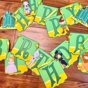 May include: A green and yellow banner with the word "Happy" and the word "Birthday" spelled out. Each letter is decorated with a character from the Wizard of Oz, including Dorothy, the Tin Man, the Cowardly Lion, the Wicked Witch of the West, and the Scarecrow.