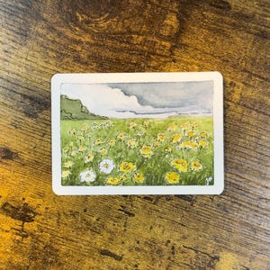 May include: Watercolor painting of a field of yellow and white flowers under a cloudy sky. The artwork is on a rectangular white card. A green hill is visible on the left side of the painting.